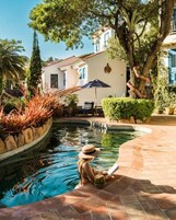 Un área de piscina con borde curvo, un patio con mesa y sombrilla, y un edificio con fachada blanca y detalles azules al fondo.