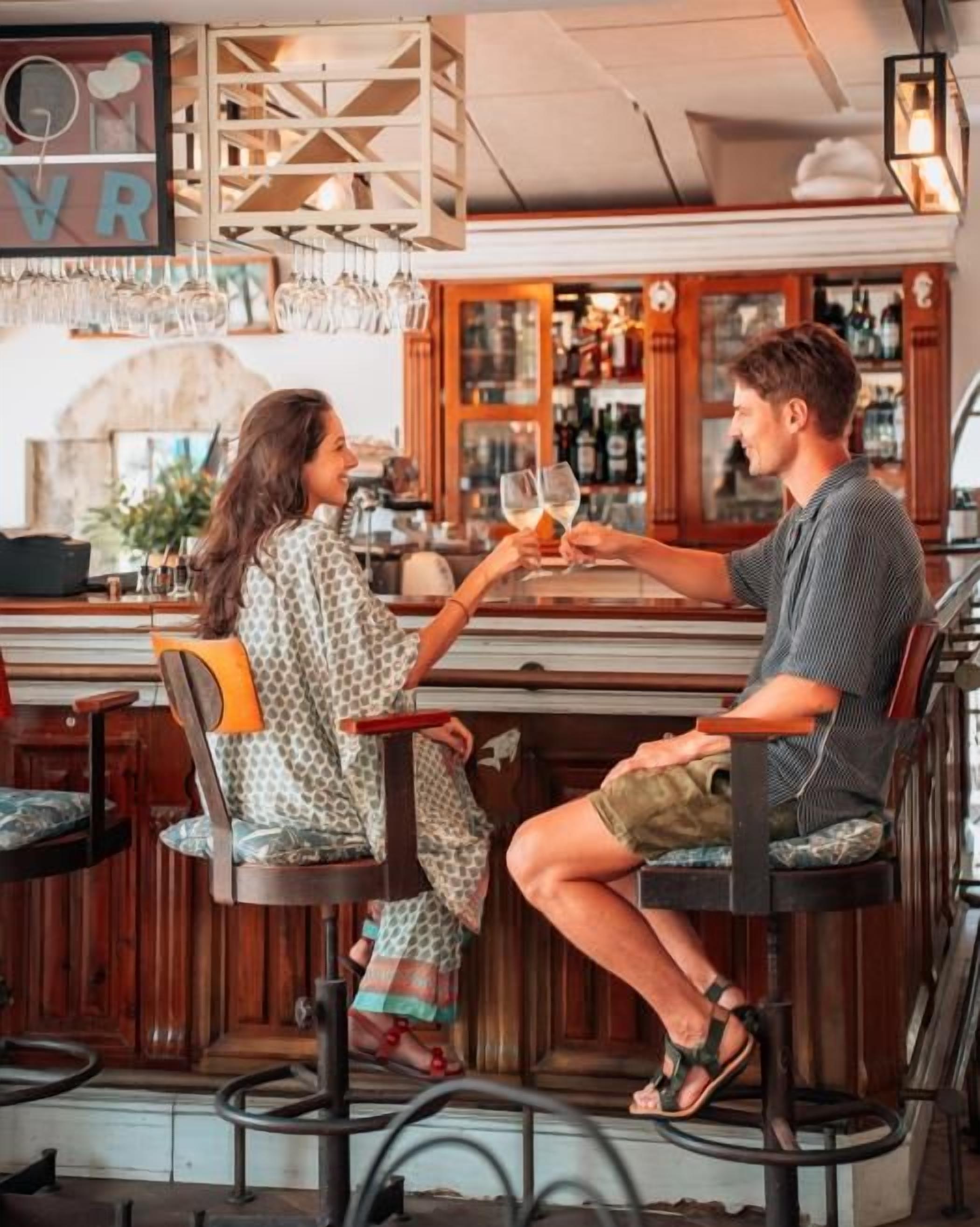 Ambiente de bar com duas pessoas sentadas no balcão, interior em madeira e adega visível.