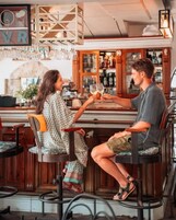 A bar setting with two people seated at a bar counter, a wooden interior, and a visible wine rack.