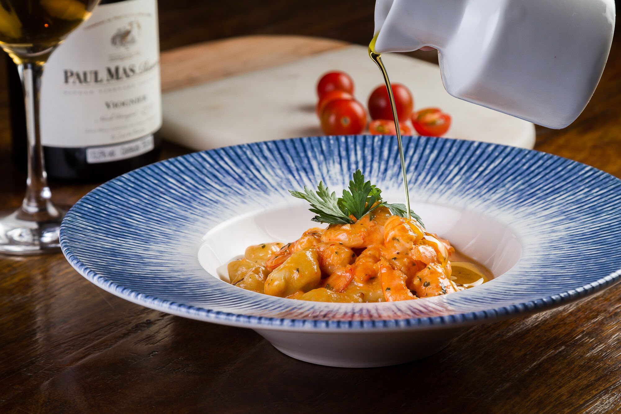 Un plato de camarones en un tazón de borde azul, adornado con perejil, con una botella de vino Paul Mas al fondo.