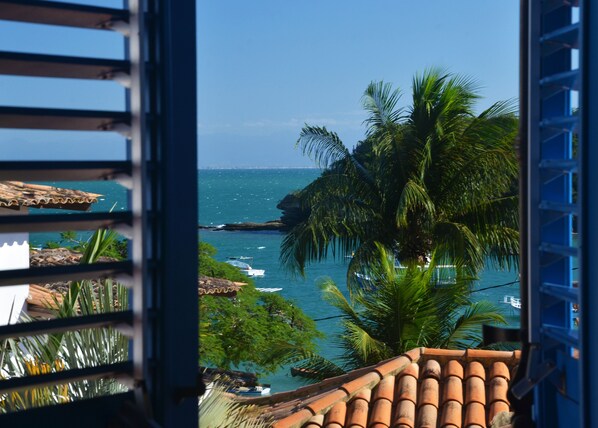 Vista costera desde un balcón con techo de tejas, palmeras y barcos en el agua.