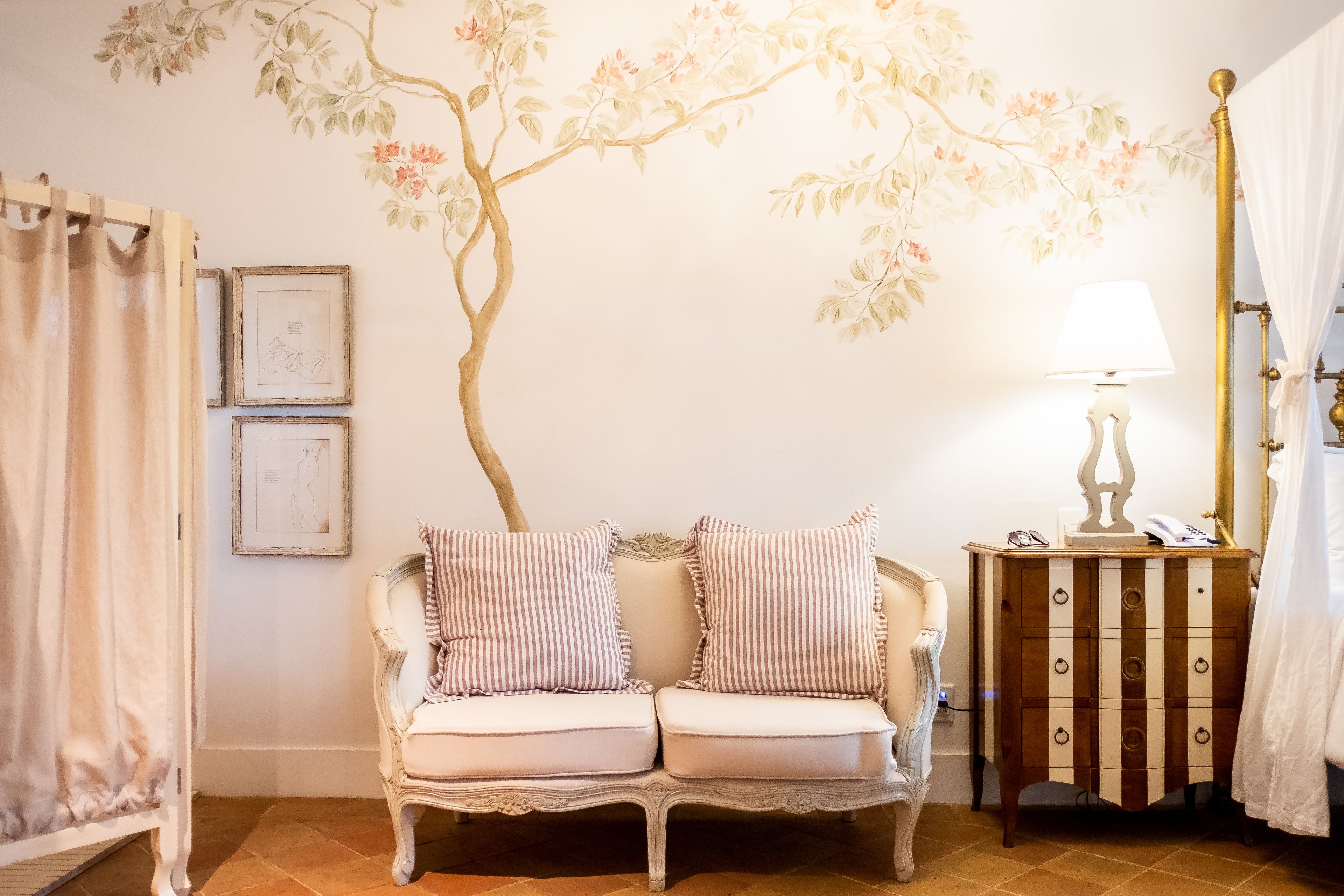 Una habitación con sofá blanco, almohadones a rayas, una mesita de noche de madera con lámpara y un mural en la pared.