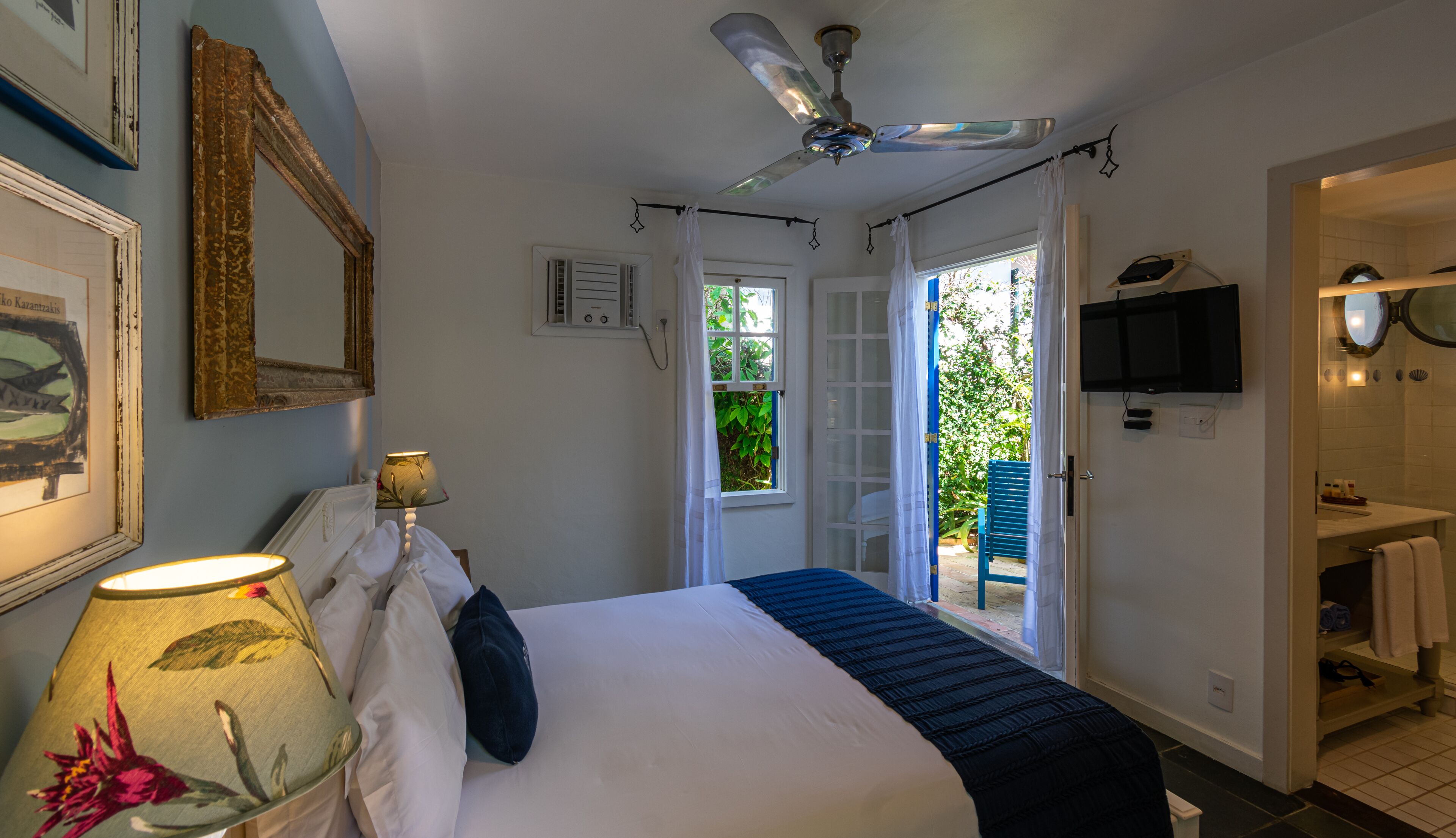 A bedroom with a bed, a ceiling fan, a TV, and a bathroom visible through an open door.