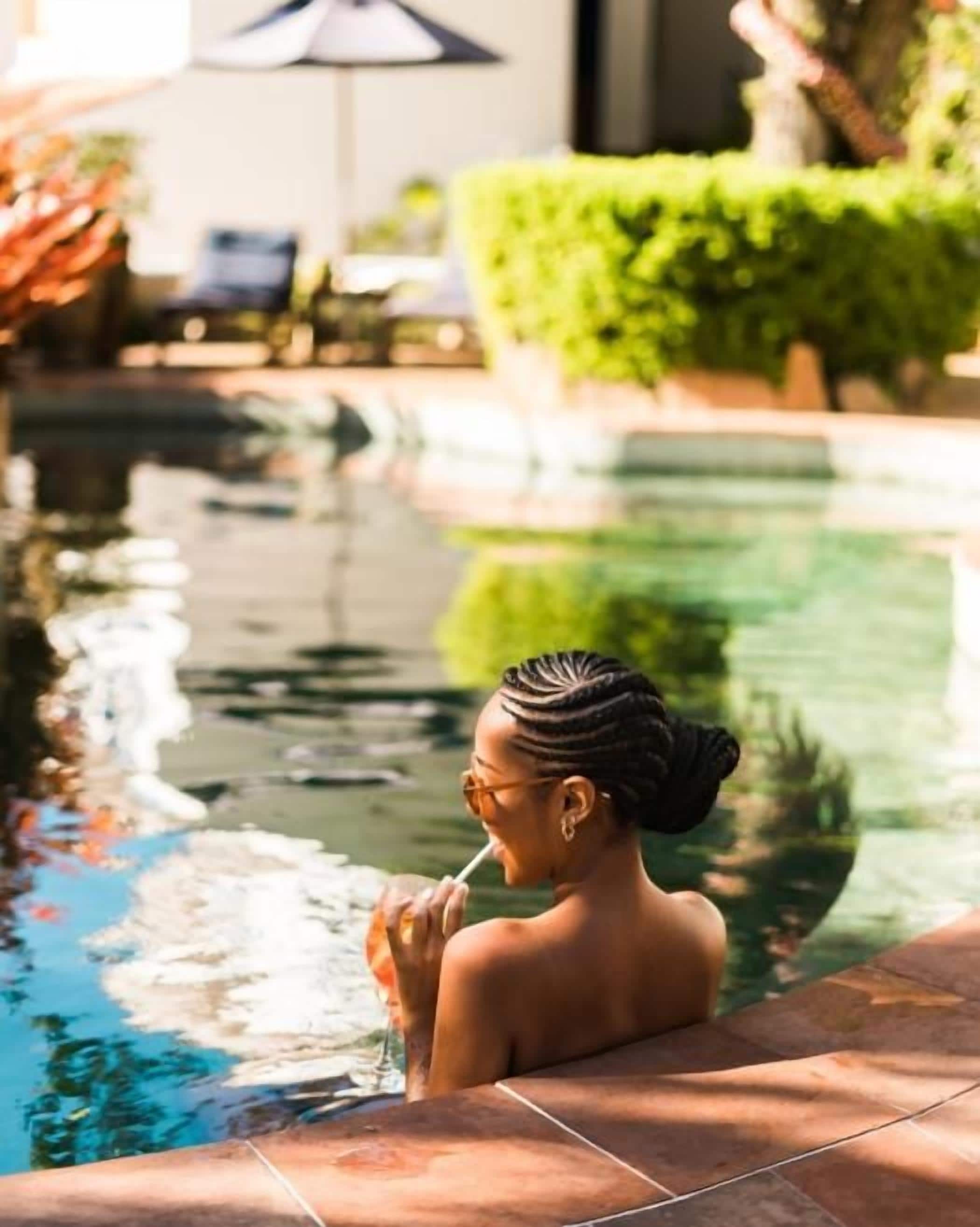 Área de piscina com uma pessoa sentada na borda, uma cadeira e uma mesa com guarda-sol.
