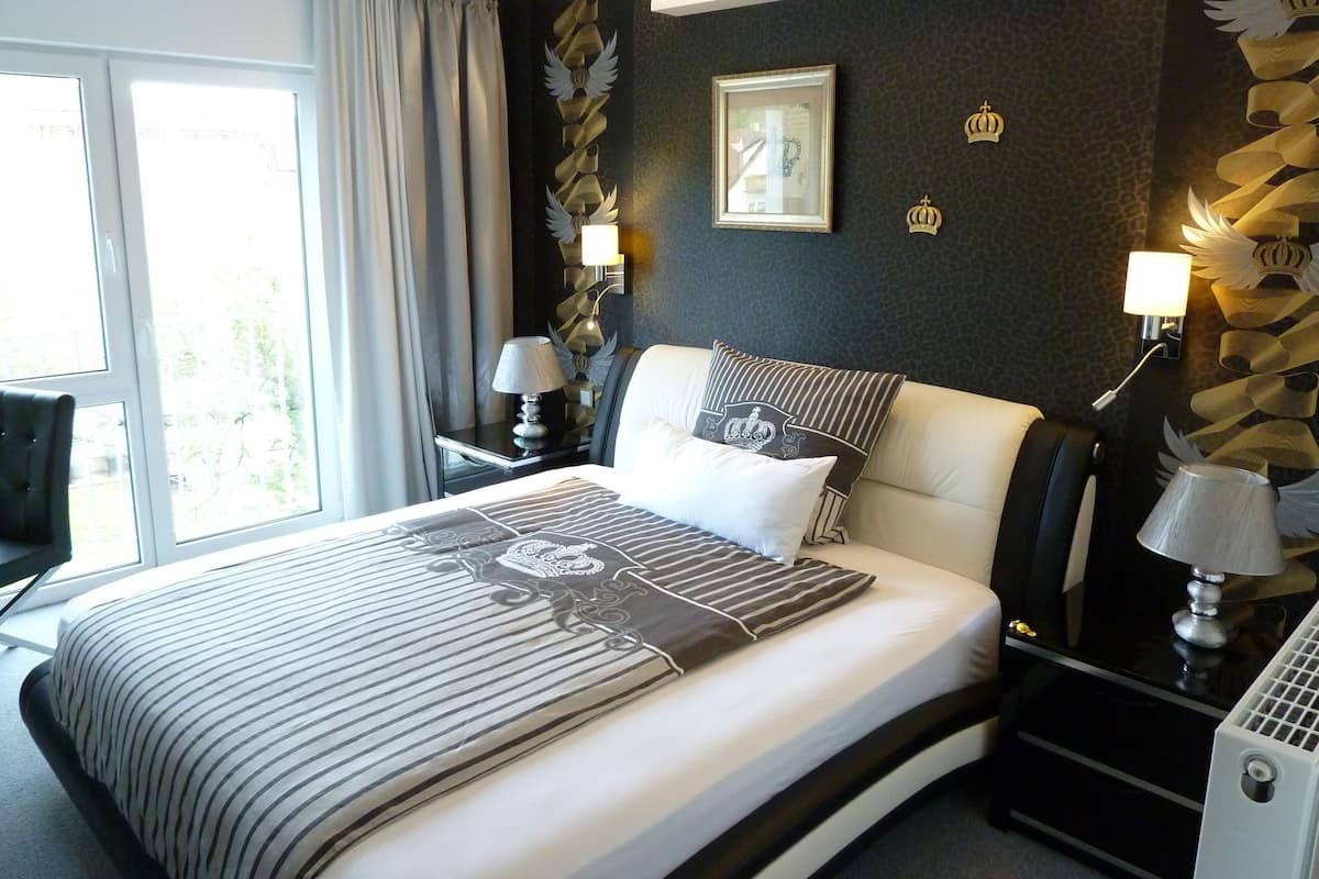 A hotel room with a large bed, a desk, and a chair. There is a window with curtains, a wall with a decorative design, and a picture frame on the wall.