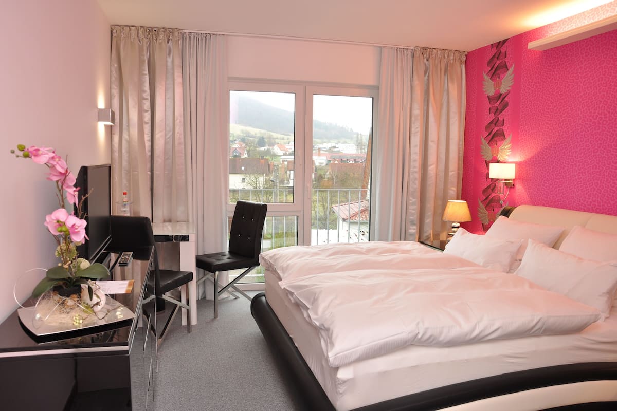A hotel room with a bed, a desk with a chair, a desk lamp, and a view of a town through large windows.