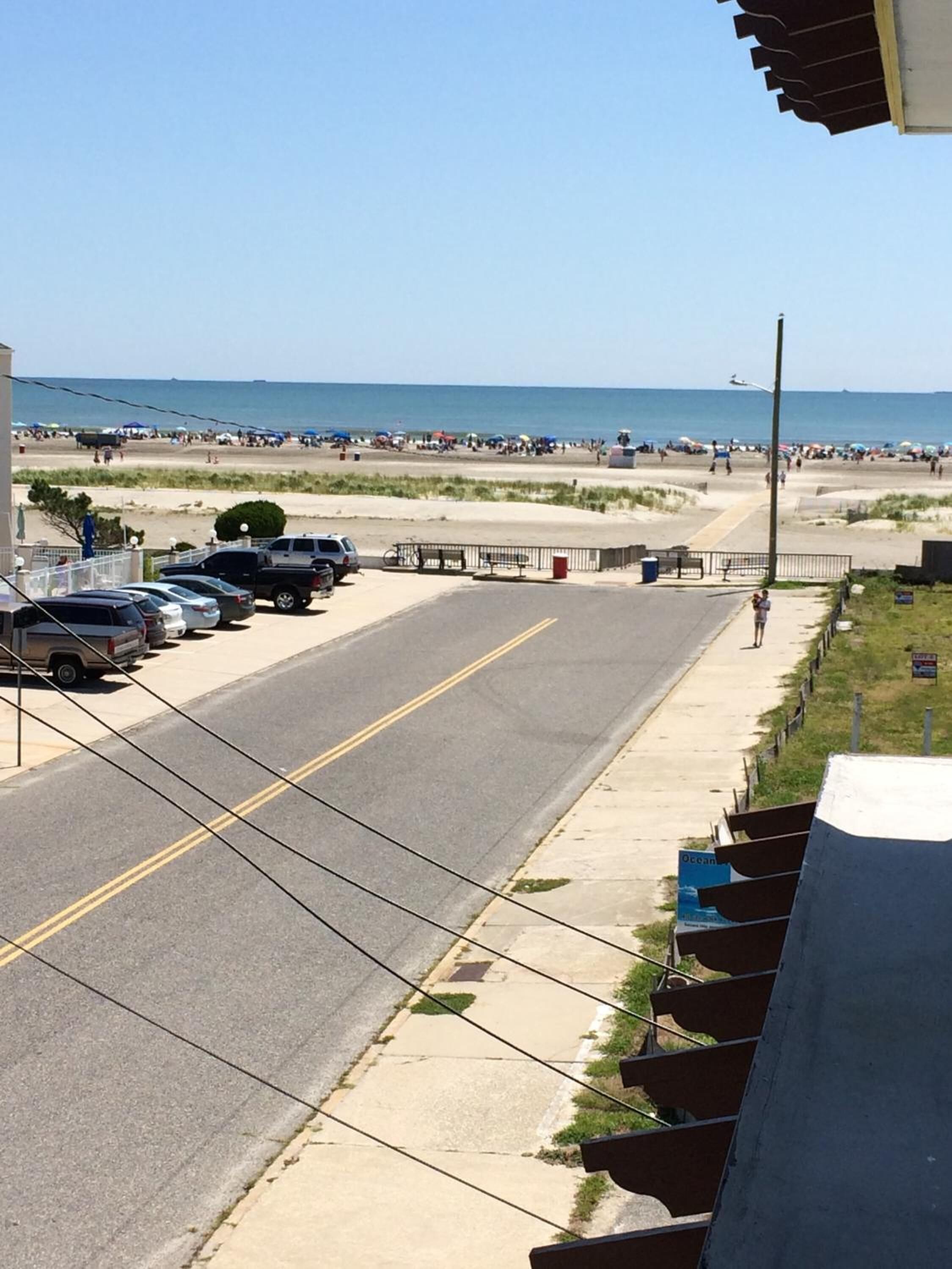 Une vue d’une rue avec des voitures garées, un trottoir et une plage en arrière-plan.