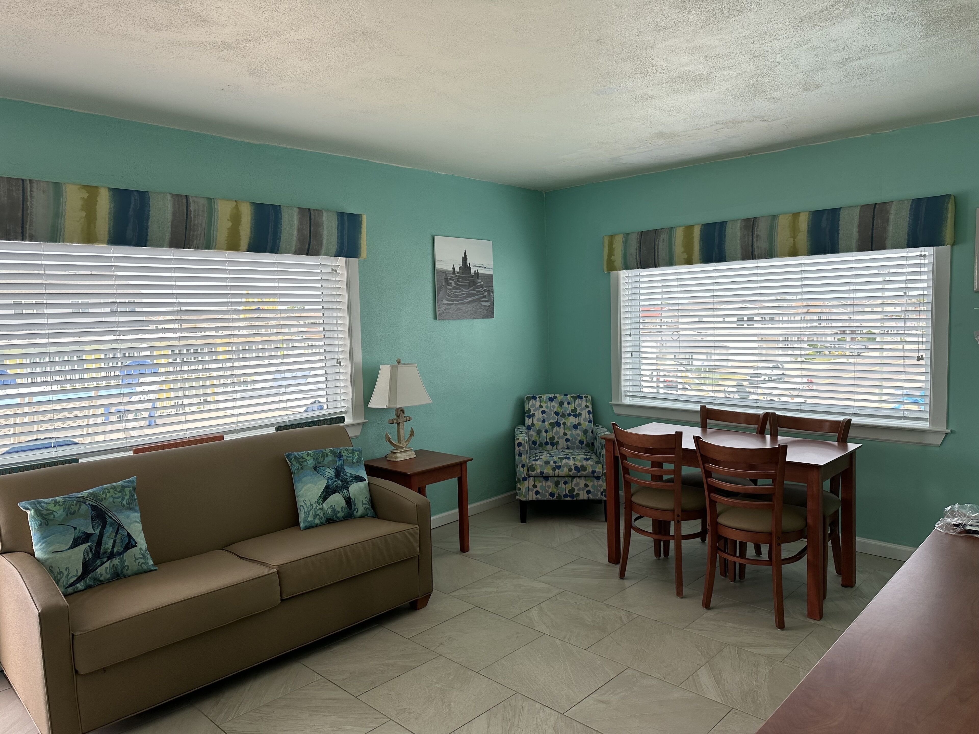 A living room with a sofa, a dining table with chairs, a patterned armchair, a lamp, and two windows with blinds.
