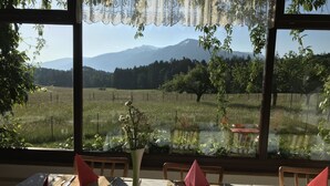 Un área de comedor con mesas dispuestas para dos, un florero con flores y vistas a montañas y campos a través de grandes ventanales.