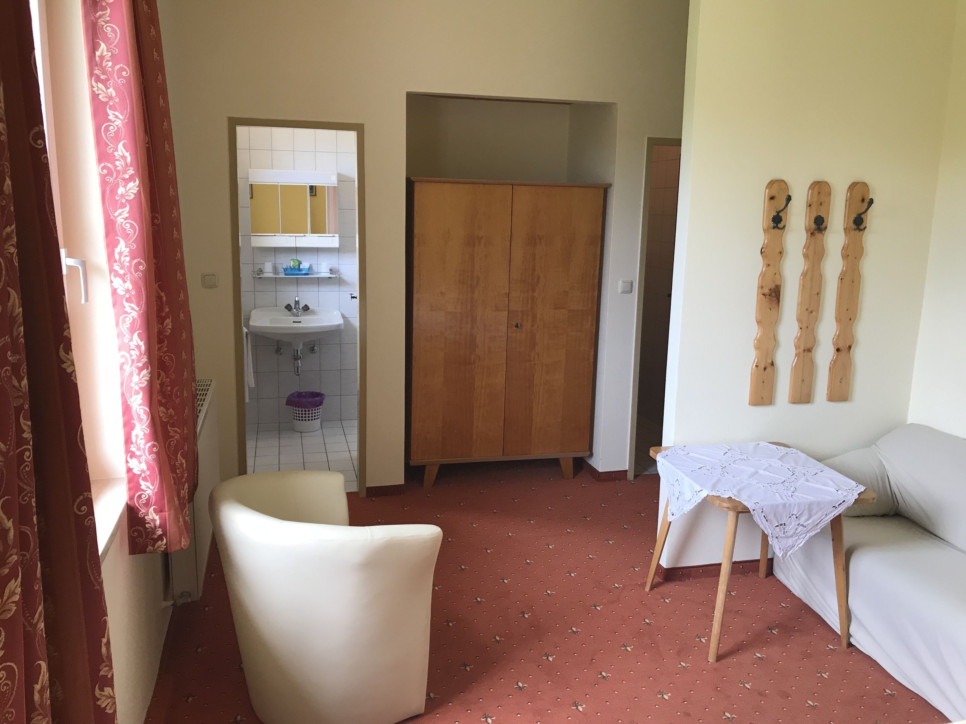 Ein Zimmer mit einem Holfischschrank, einem weißen Stuhl, einem kleinen Tisch mit Spitzenbettdecke und einem Bett mit weißer Laken.