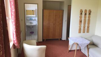 Ein Zimmer mit einem Holfischschrank, einem weißen Stuhl, einem kleinen Tisch mit Spitzenbettdecke und einem Bett mit weißer Laken.