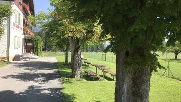 A pathway leading to a building with a white facade and green shutters, surrounded by trees and picnic tables in a grassy area.