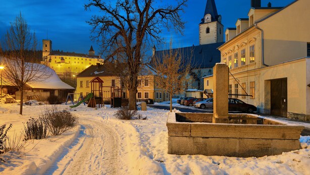A snow-covered street with a castle in the background, a church steeple, and residential buildings.