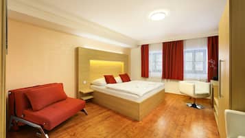 A modern hotel room with a wooden floor, a bed with red pillows, a red sofa, and a hairdressing chair.