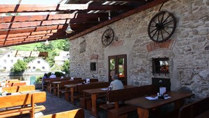 An outdoor seating area with wooden benches and tables, stone walls, and a view of a river and buildings in the background.