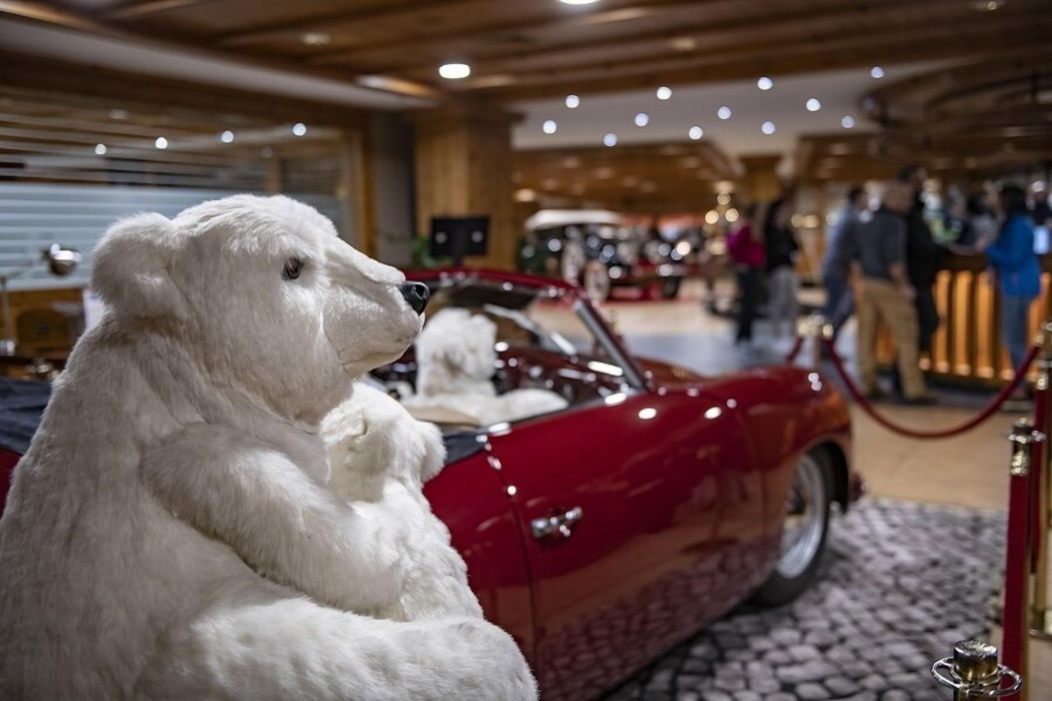 Une statue d’un ours polaire en peluche, placée à côté d’une voiture rouge décapotable dans un musée, avec des expositions et des visiteurs.