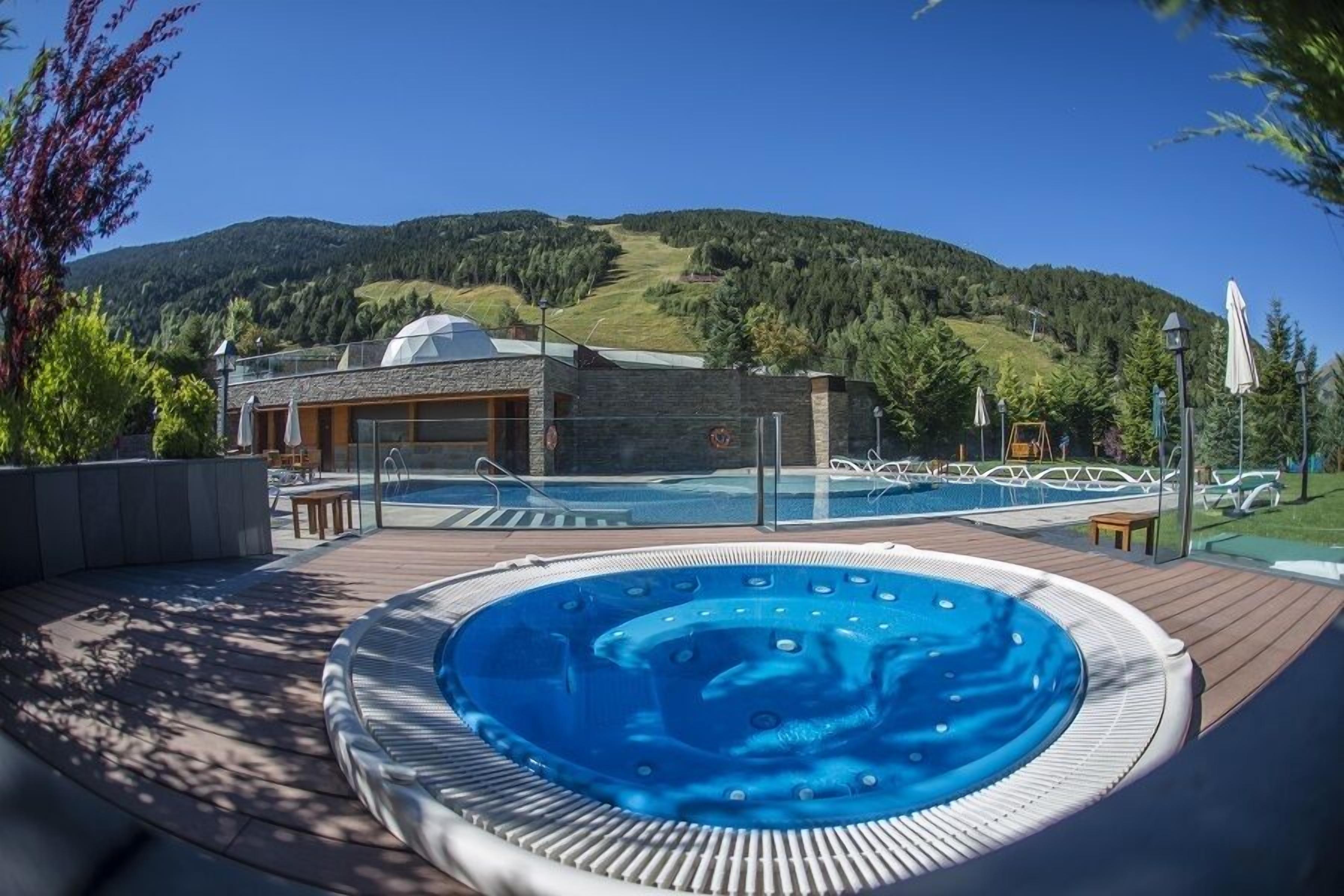 A hot tub on a wooden deck with a pool in the background, a building with a dome, and a mountainous landscape.