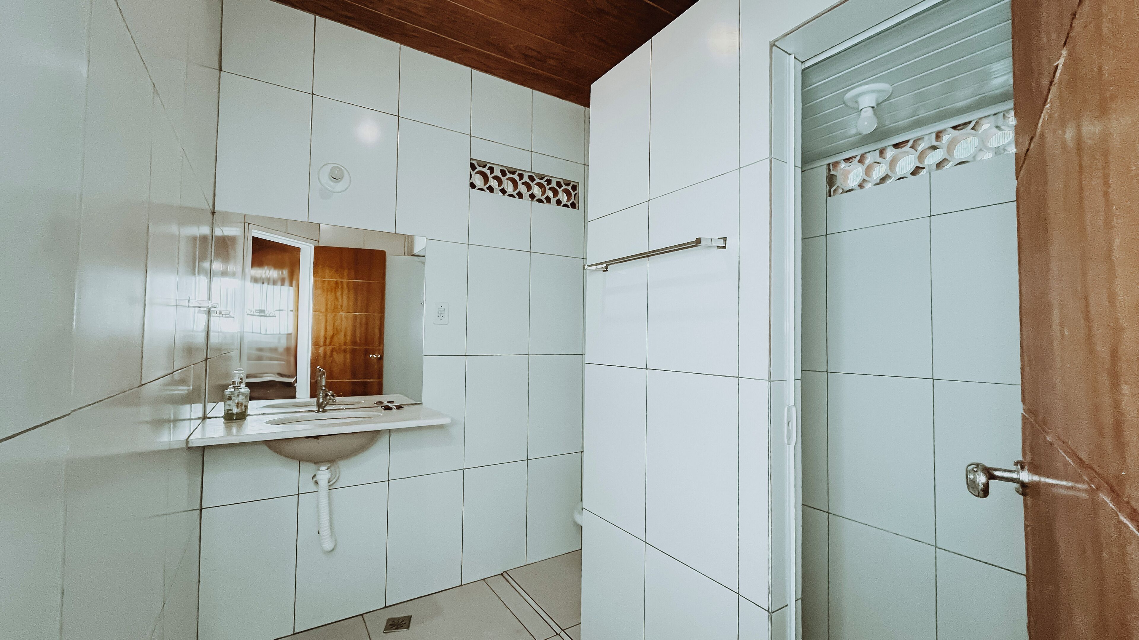 A bathroom with white tiles, a wooden ceiling, a sink with a mirror, and a shower area.