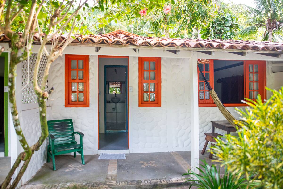 Um pequeno edifício térreo com telhado de telhas e fachada branca. Há um banco verde na parte externa e uma pequena mesa no interior.