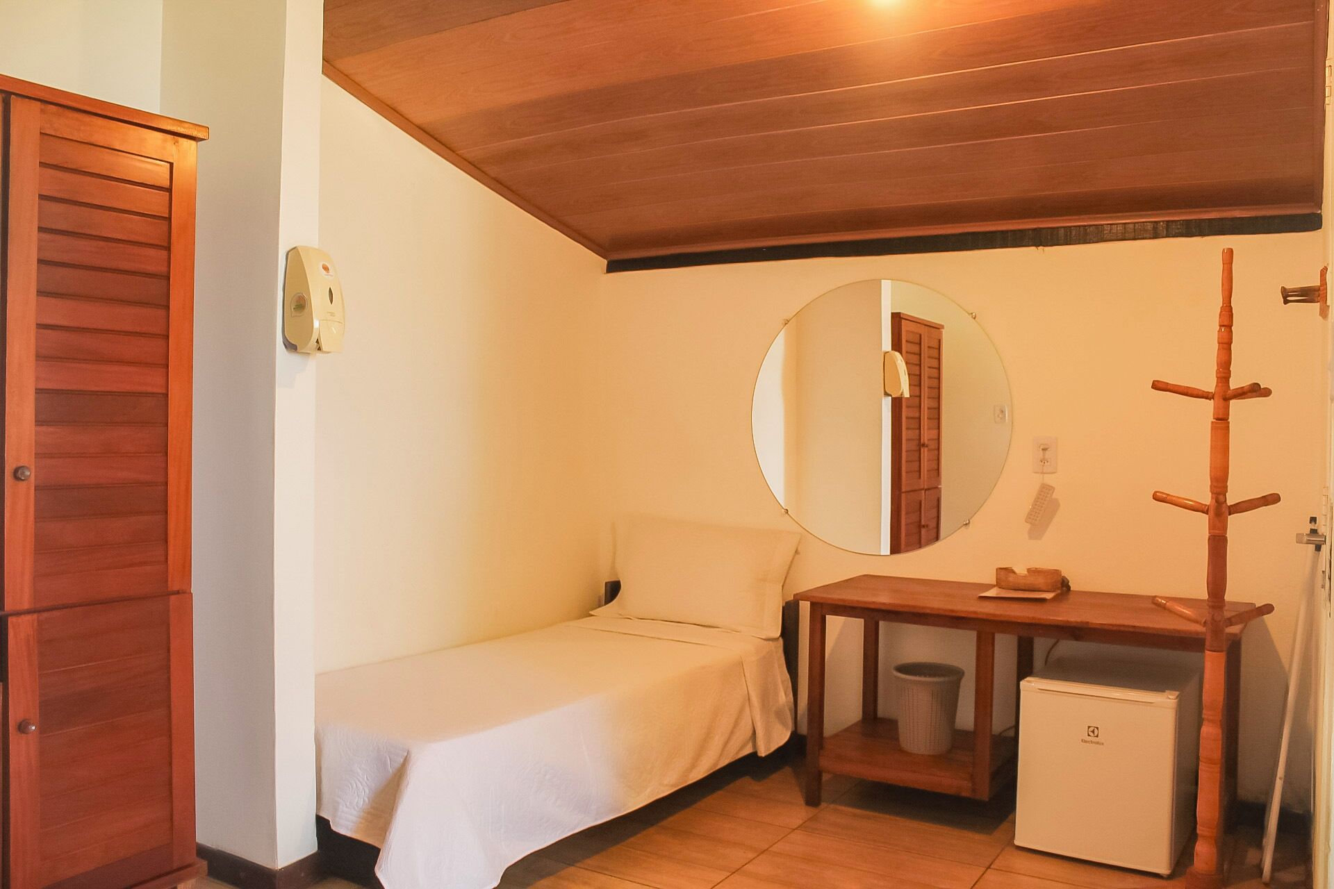 Ein kleiner, gemütlicher Raum mit einem Einzelbett, einem Holzschrank, einem runden Spiegel, einem kleinen Tisch mit Hocker und einem kompakten Kühlschrank.