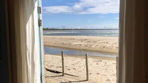 Vista desde una ventana a una playa con orilla arenosa, una valla de madera y cielo despejado.