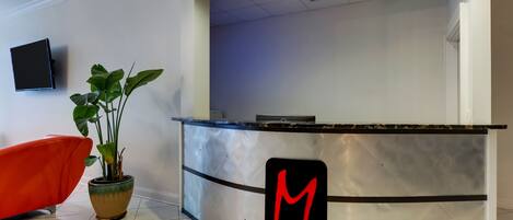A reception desk with a 'M' logo, a potted plant, and a red chair.