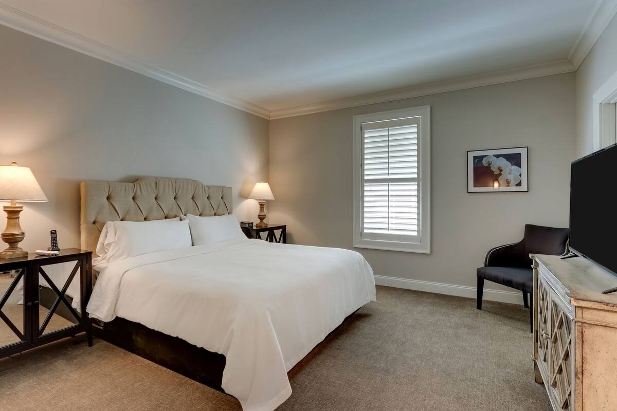 A neatly arranged bedroom with a large bed, two bedside tables with lamps, a chair, and a TV on a wooden cabinet.