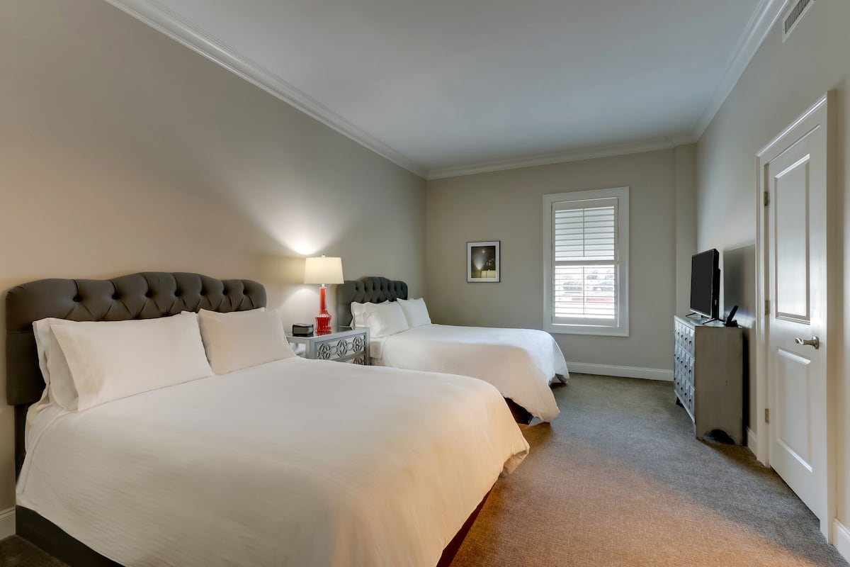 A hotel room with two beds, a television, a lamp, and a window with blinds.