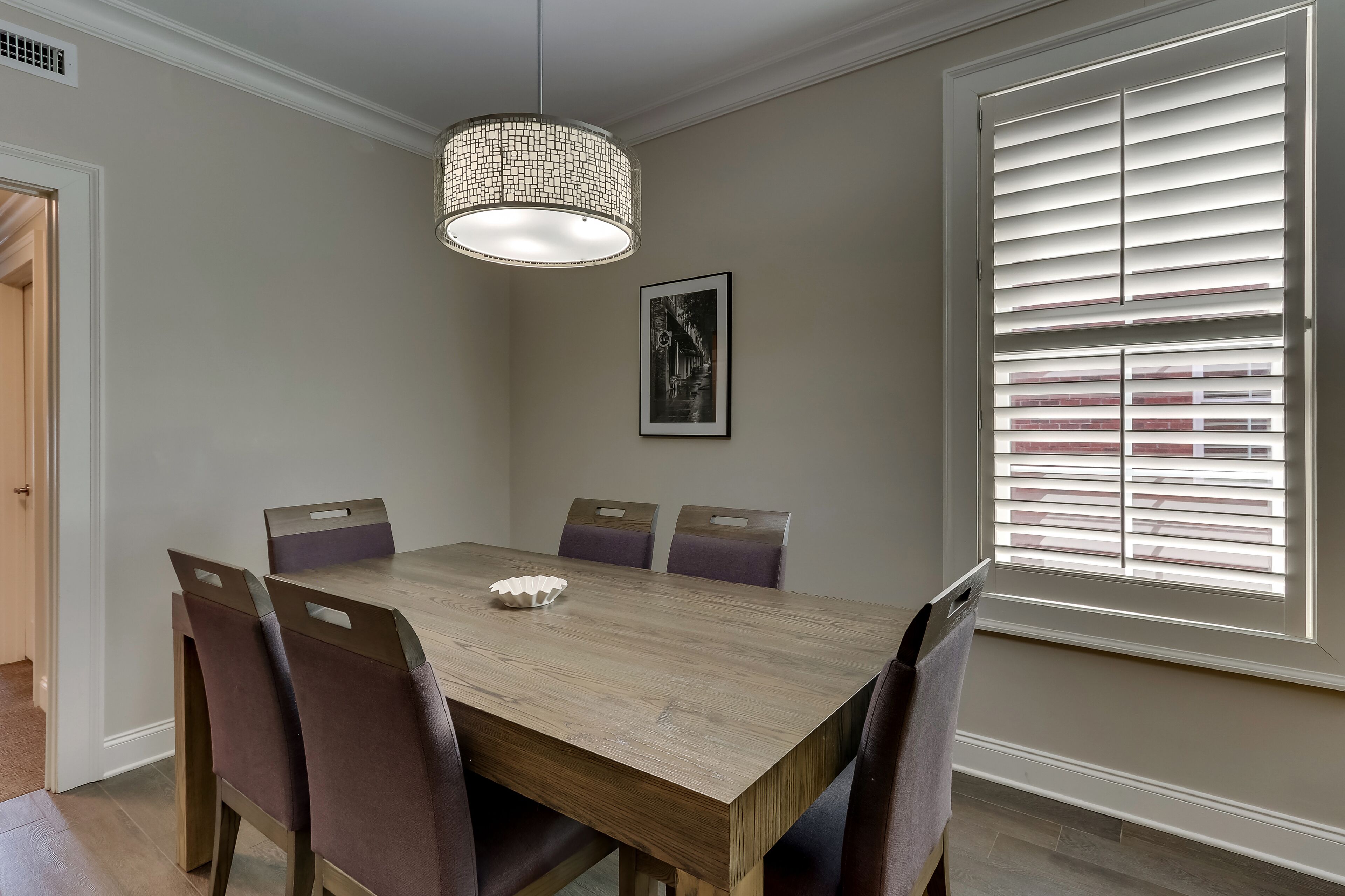 A dining room with a wooden table, six chairs, a pendant light, a window with blinds, and a framed picture on the wall.