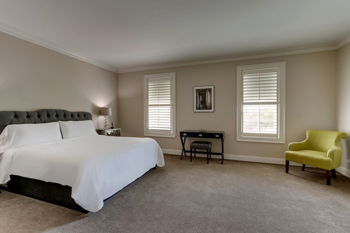 A bedroom with a bed, desk, chair, and two windows with blinds.