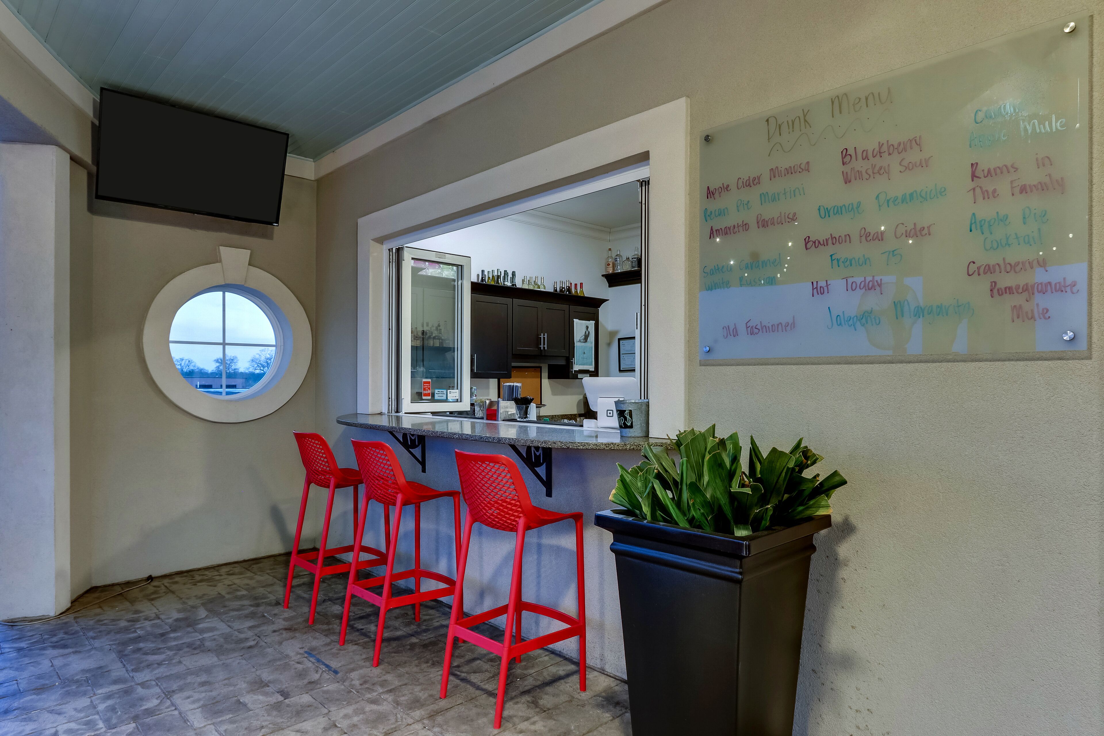 Un espace bar avec un menu de boissons, des tabourets rouges, une grande fenêtre et une plante en pot.