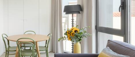 A modern living room with a round table, green chairs, a sofa with a patterned pillow, and a vase of sunflowers by a window.