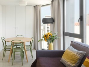 A modern living room with a round table, green chairs, a sofa with a patterned pillow, and a vase of sunflowers by a window.