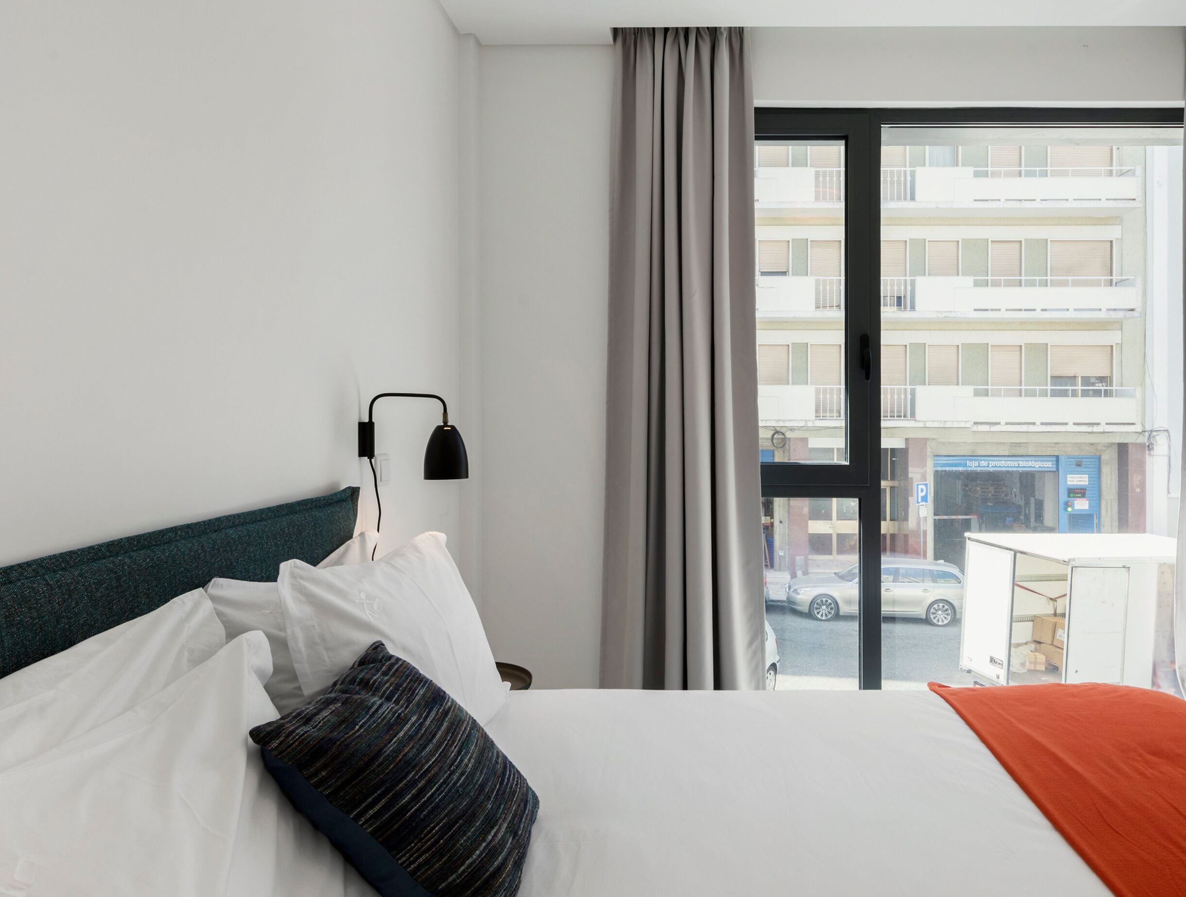 A hotel room with a bed, a nightstand, a lamp, a window with a view of the street, and a curtain.