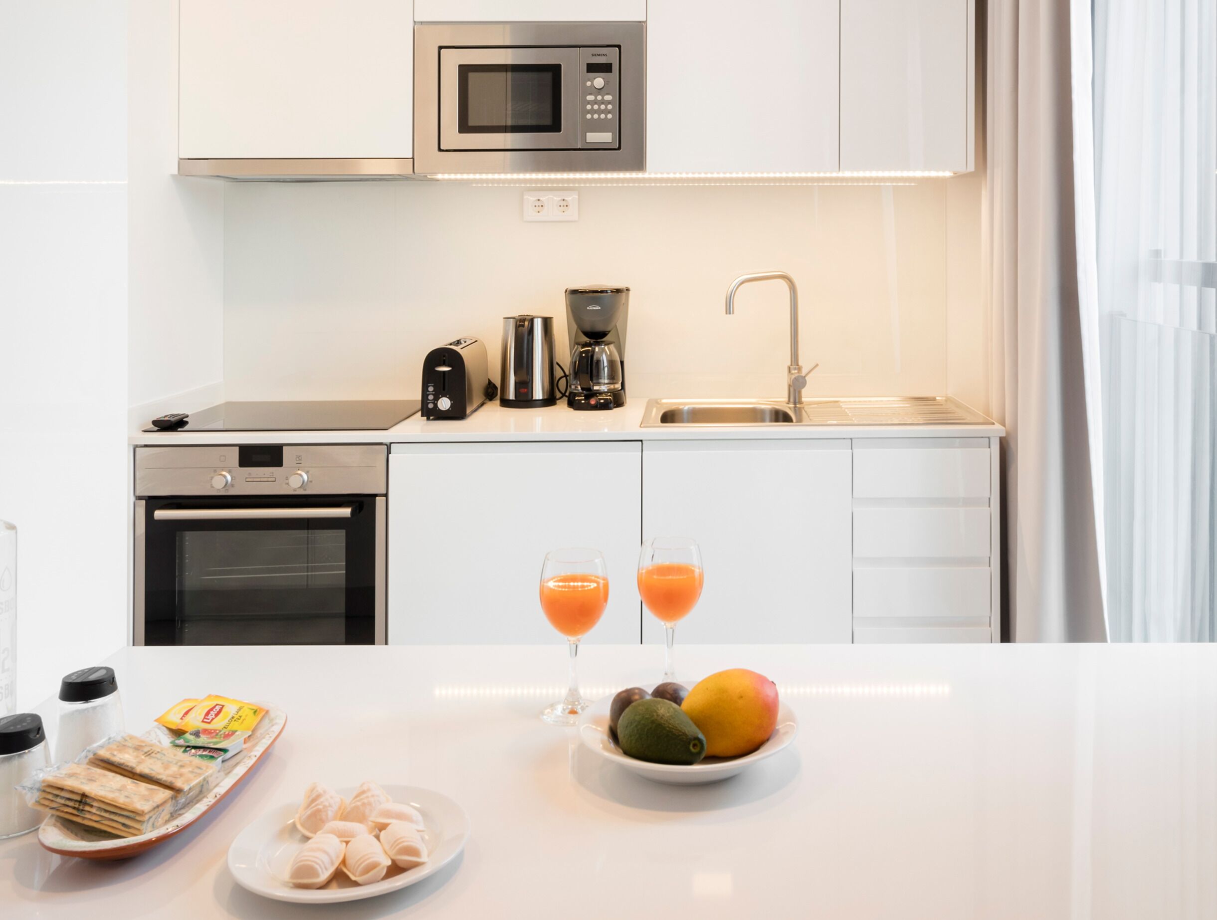 Una cocina moderna con microondas, tostadora, cafetera y pileta. Hay dos vasos de jugo de naranja y un bol de fruta en la encimera.