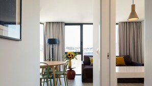 Una sala de estar moderna con un sofá, un comedor con mesa y sillas, una lámpara y vistas amplias a través de grandes ventanales.