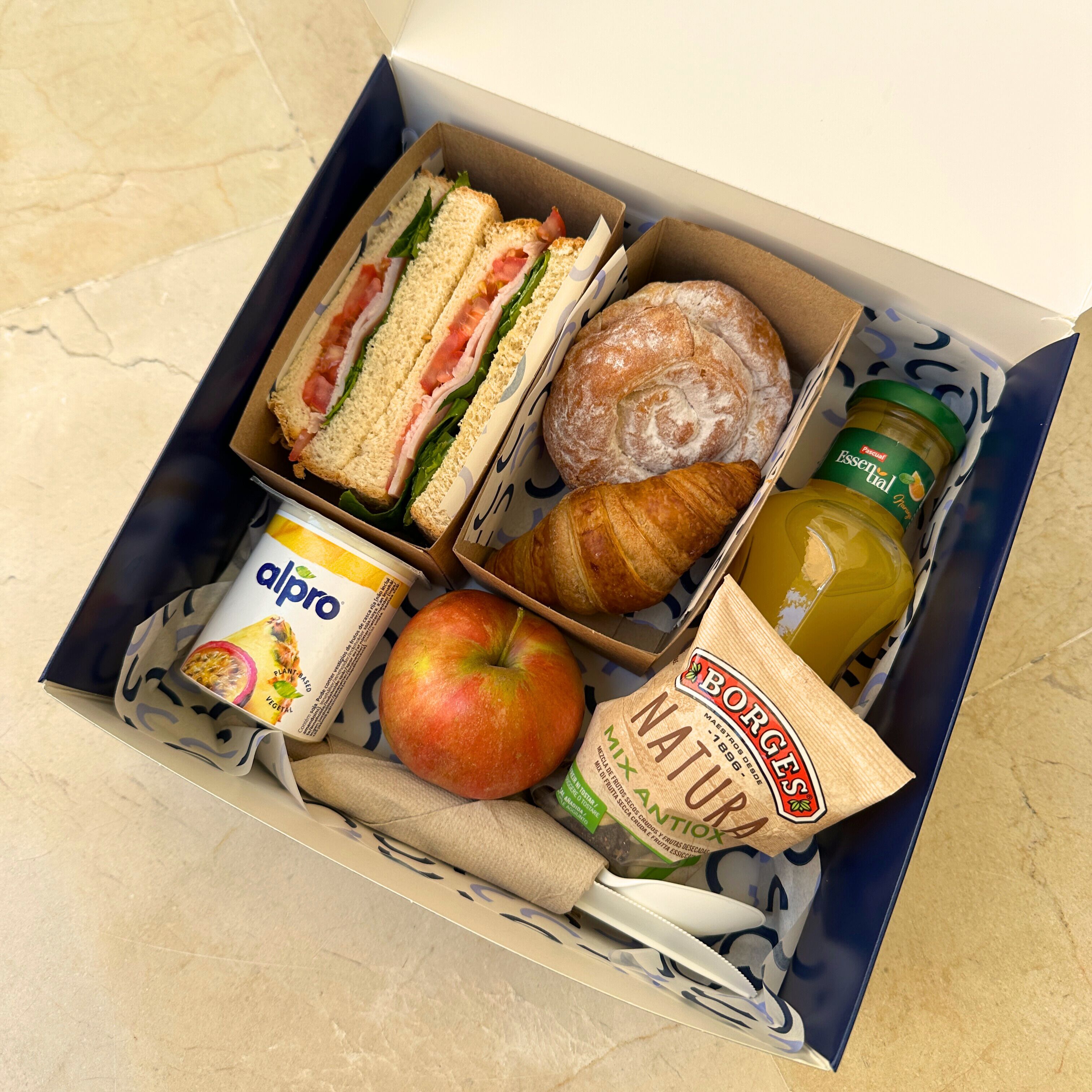 A lunch box containing sandwiches, a croissant, an apple, a bottle of juice, a yogurt container, and a bag of bread.