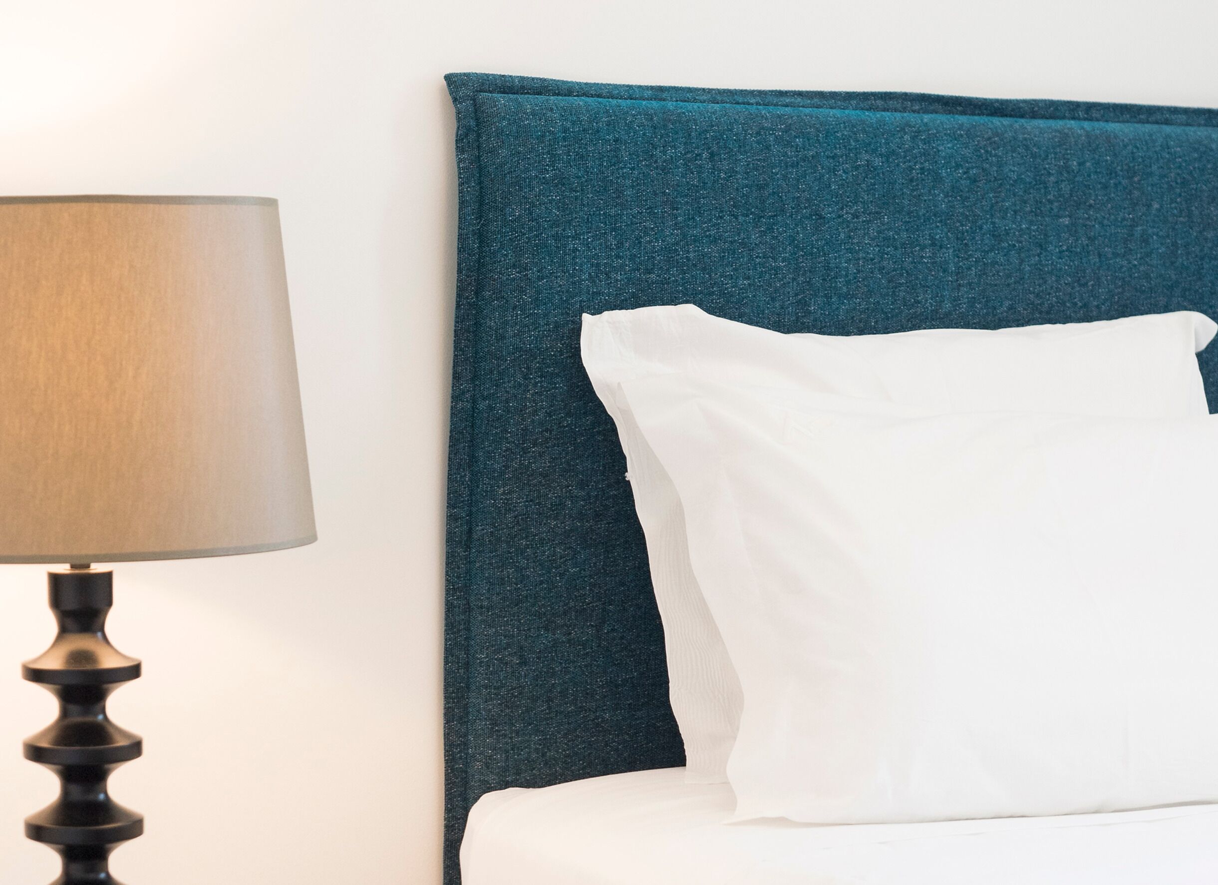 A bed with a teal headboard, a white pillow, and a lamp with a beige shade.