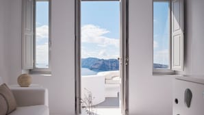 A minimalist living room with a white sofa, a coffee table with a book and a vase, and a view of the sea and mountains through open doors.