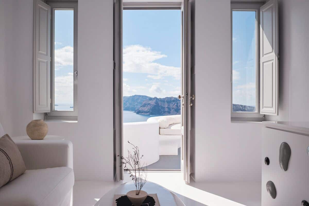 A minimalist living room with a white sofa, a coffee table with a book and a vase, and a view of the sea and mountains through open doors.