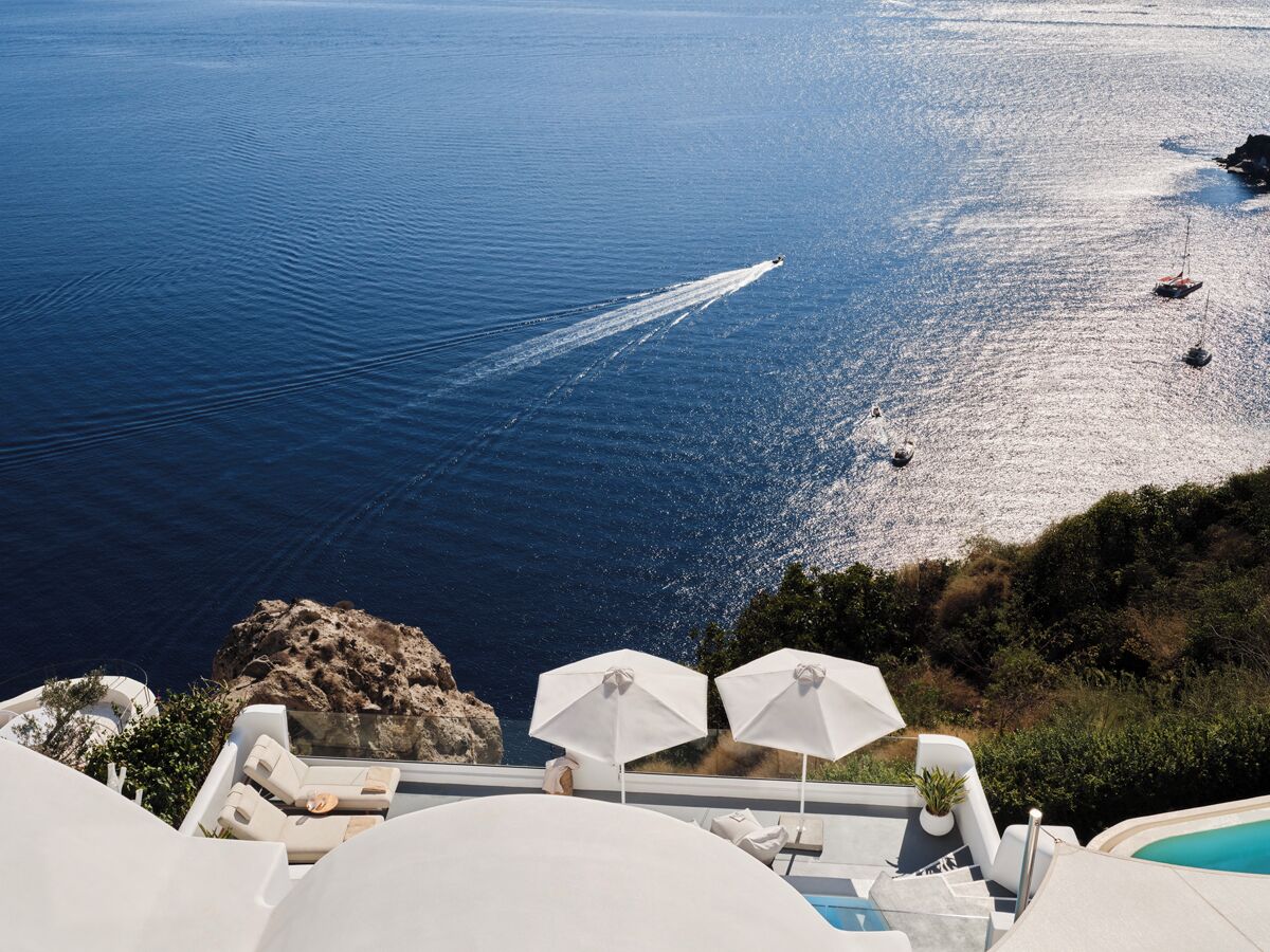 Une vue sur la mer avec un bateau laissant une vague derrière lui, un promontoire rocheux et un espace piscine avec des chaises longues et des parasols.
