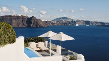 Zona junto a la piscina con mobiliario blanco y sombrillas, con vistas a un cuerpo de agua y montañas a lo lejos.