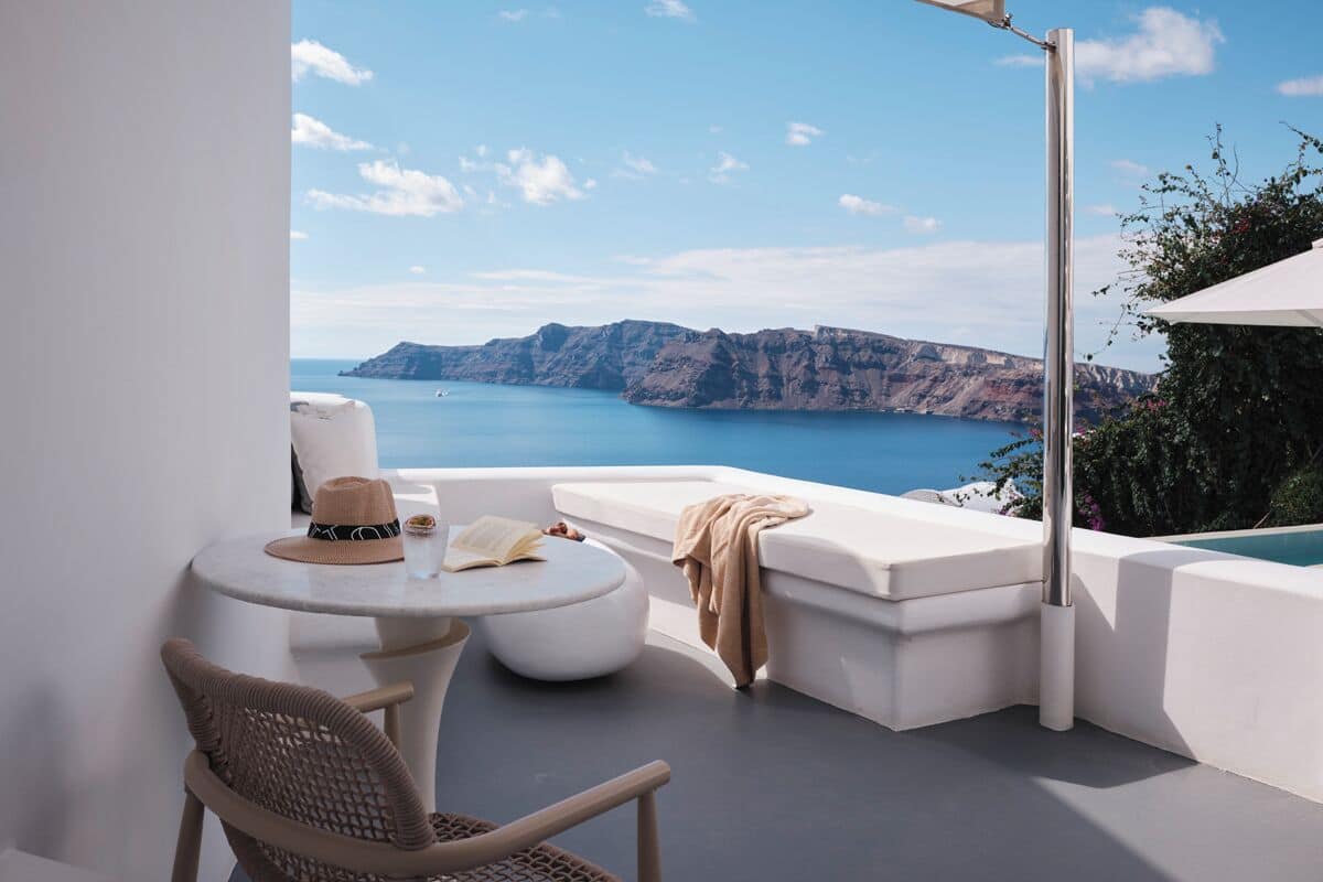 A balcony with a white cushioned seating area, a round table with a hat and book, and a view of the sea and cliffs.