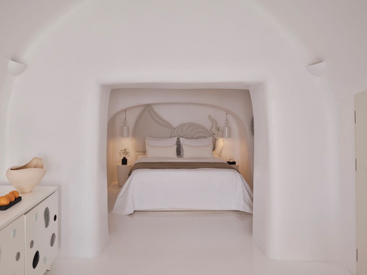 A minimalist bedroom with a bed centered under a curved ceiling, flanked by white walls and a white dresser with a bowl on top.