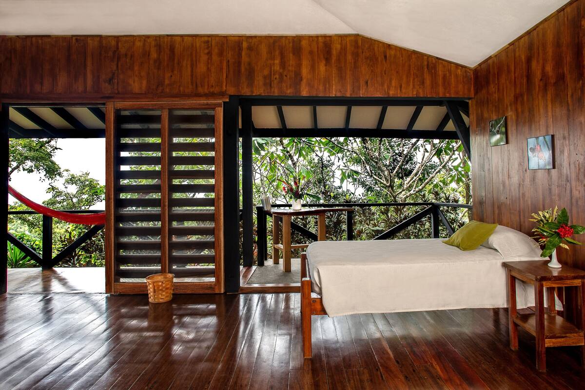 A wooden room with a bed, a small table, and a view of greenery outside.