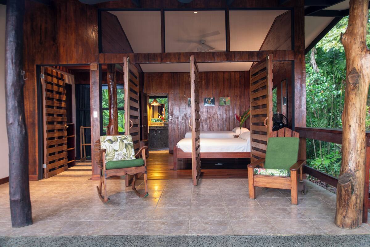 A wooden room with a bed, two chairs, and a view of greenery outside.
