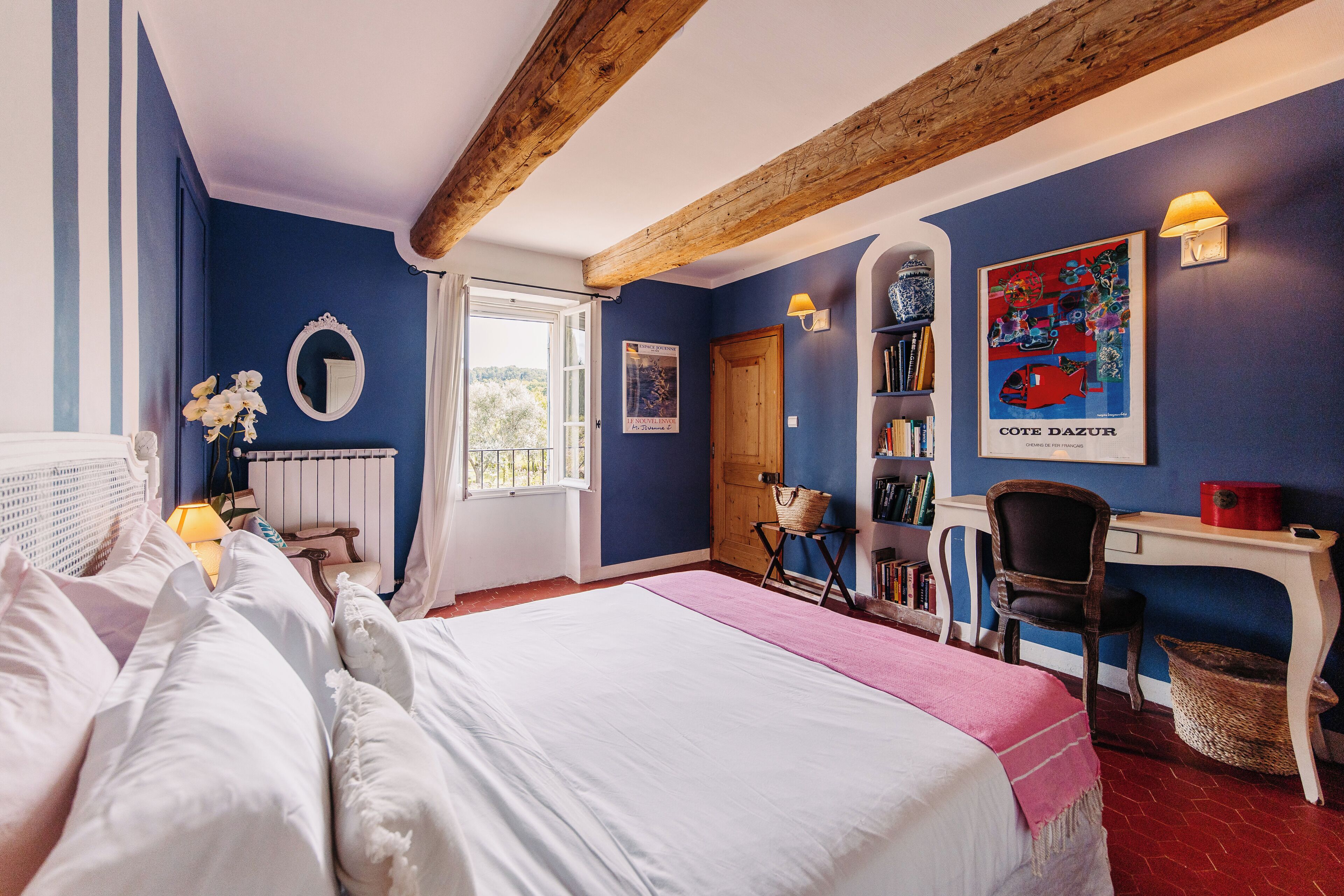A bedroom with a large bed, a desk, a chair, a bookshelf, and a window with a view of greenery.