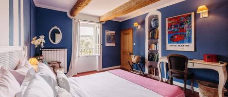 A bedroom with a large bed, a desk, a chair, a bookshelf, and a window with a view of greenery.