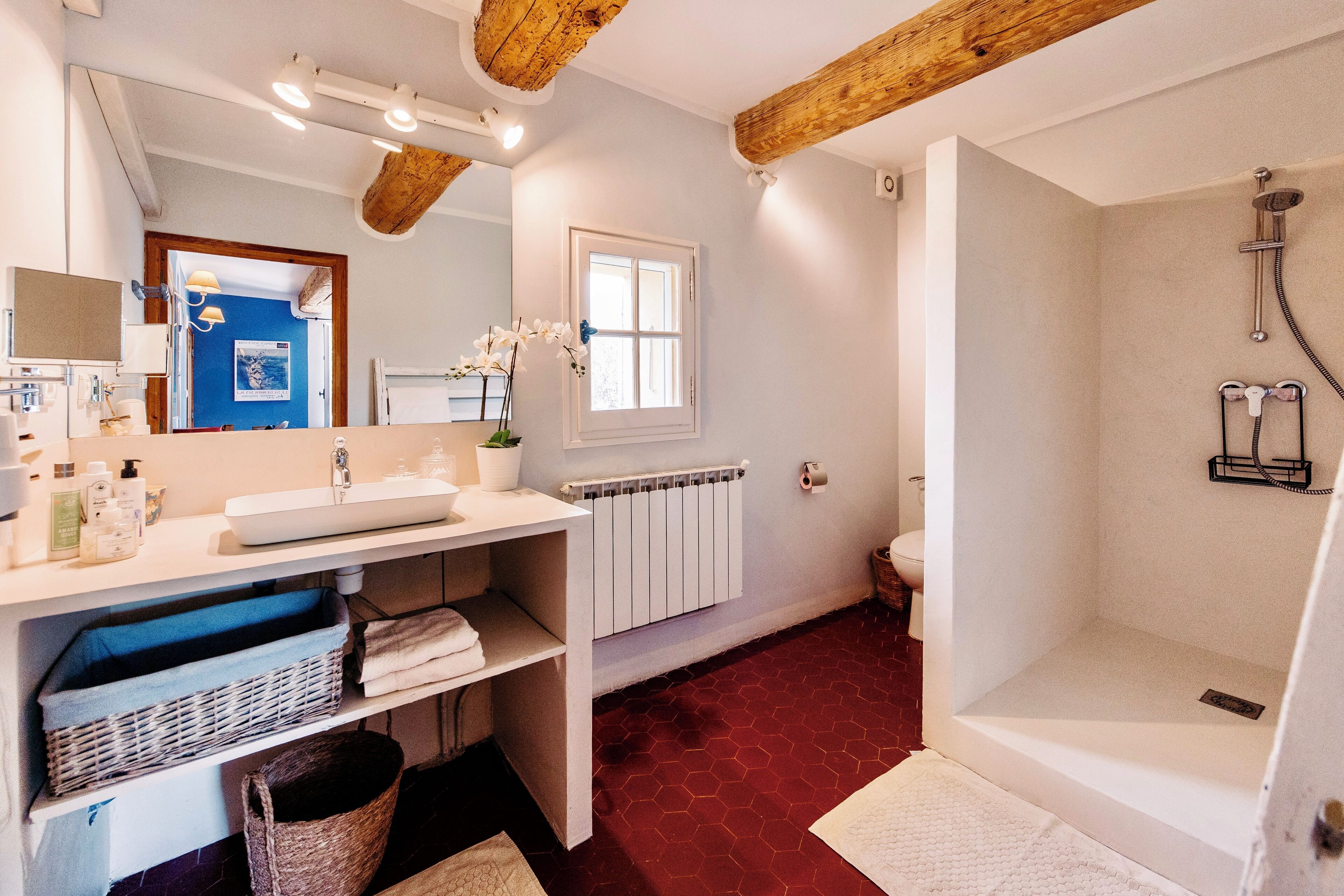 Une salle de bain avec un lavabo blanc, un miroir et une douche. Il y a une fenêtre au-dessus du lavabo et un radiateur en dessous.