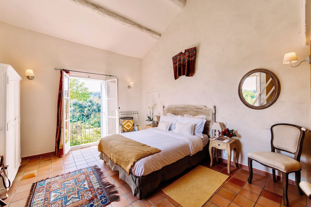 A bedroom with a bed, a chair, a nightstand, a mirror, and a view of the outdoors.